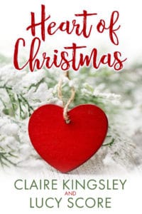 The book cover for Heart of Christmas, a bonus story from Claire Kingsley and Lucy Score, is an image of a wooden red heart on a piece of twine on a bed of pine leaves covered in snow.