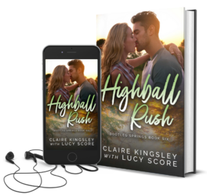 The book cover for Highball Rush, a novel by Claire Kingsley with Lucy Score, is an image of a blonde woman leaning in to kiss a dark haired man while the sun sets in the background, she has one arm wrapped around his shoulders. Next to the book is a phone showing the cover with a pair of wired earbuds.