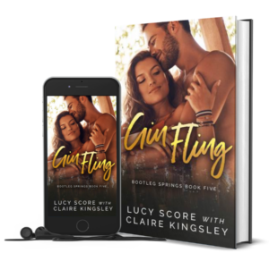 The book cover of Gin Fling, a novel by Lucy Score with Claire Kingsley, is an image of a dark haired woman wrapped in the arms of a bearded man with her back to his front, his face pressed to her forehead. Next to the book is a phone showing the cover with a pair of wired earbuds.
