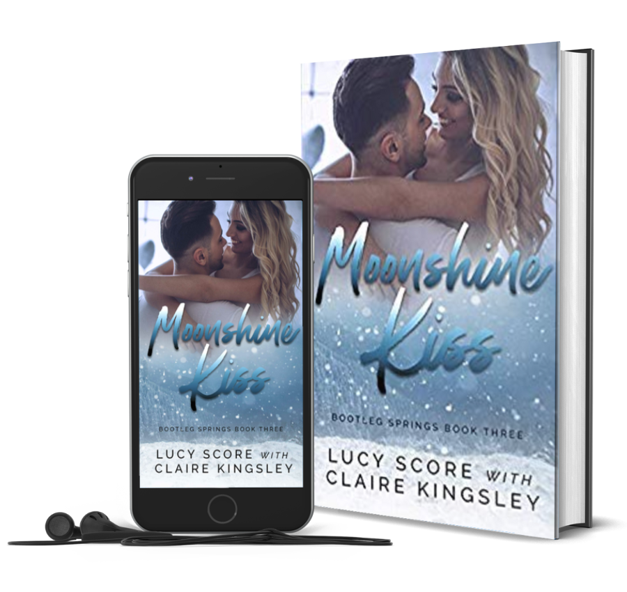 The book cover for Moonshine Kiss, a novel by Lucy Score with Claire Kingsley, is an image of a blonde woman being held by a dark haired man while she has her arms around his shoulders, their faces close while they smile at each other. Next to the book is a phone showing the cover with a pair of wired earbuds.