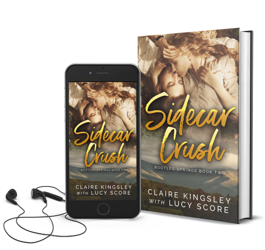 The book cover for Sidecar Crush, a novel by Claire Kingsley with Lucy Score, is an image of a man and a woman laying together in the grass their heads leaning in close for a kiss, his one hand lifted to run through her blonde curly hair. Next to the book is a phone showing the cover with a pair of wired earbuds.