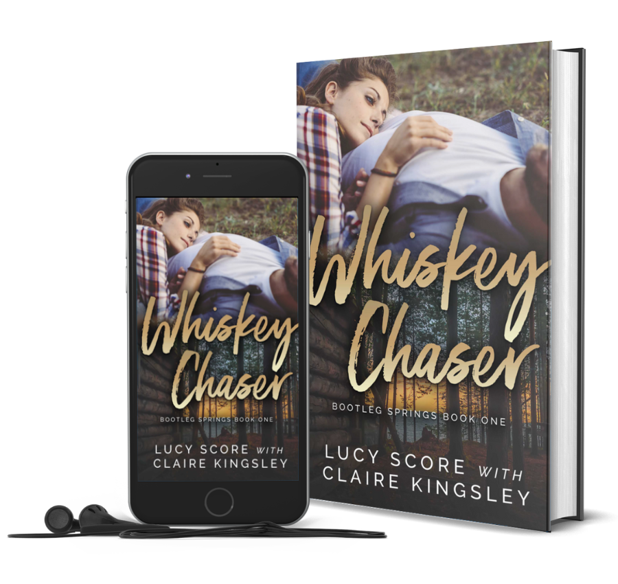 The book cover for Whiskey Chaser, a novel by Lucy Score with Claire Kingsley, is an image of a man in a white t-shirt laying in the grass while a woman with red hair rests her head on his stomach. Next to the book is a phone showing the cover with a pair of wired earbuds.