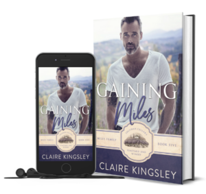 The book cover for Gaining Miles, a small-town romance by Claire Kingsley, is an image of a man in a white t-shirt with gray in his dark hair and beard, standing with a small smile on his face, a background of trees and hills behind him. There are shades of purple and white. Next to the book is a phone showing the cover with a pair of wired earbuds.