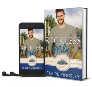 The book cover for Reckless Miles, a small-town romance by Claire Kingsley, is an image of a smiling dark haired man in a light green long sleeve shirt standing in a vineyard with grapes in the background. There are shades of blue and white. Next to the book is a phone showing the cover with a pair of wired earbuds.