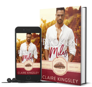 The book cover for Broken Miles, a small-town romance by Claire Kingsley, is an image of a dark haired man in a white button down shirt standing in a vineyard with the sun setting in the background. He has his hands tucked into his pockets and a serious expression on his face. There are shades of dark red and white. Next to the book is a phone showing the cover with a pair of wired earbuds.