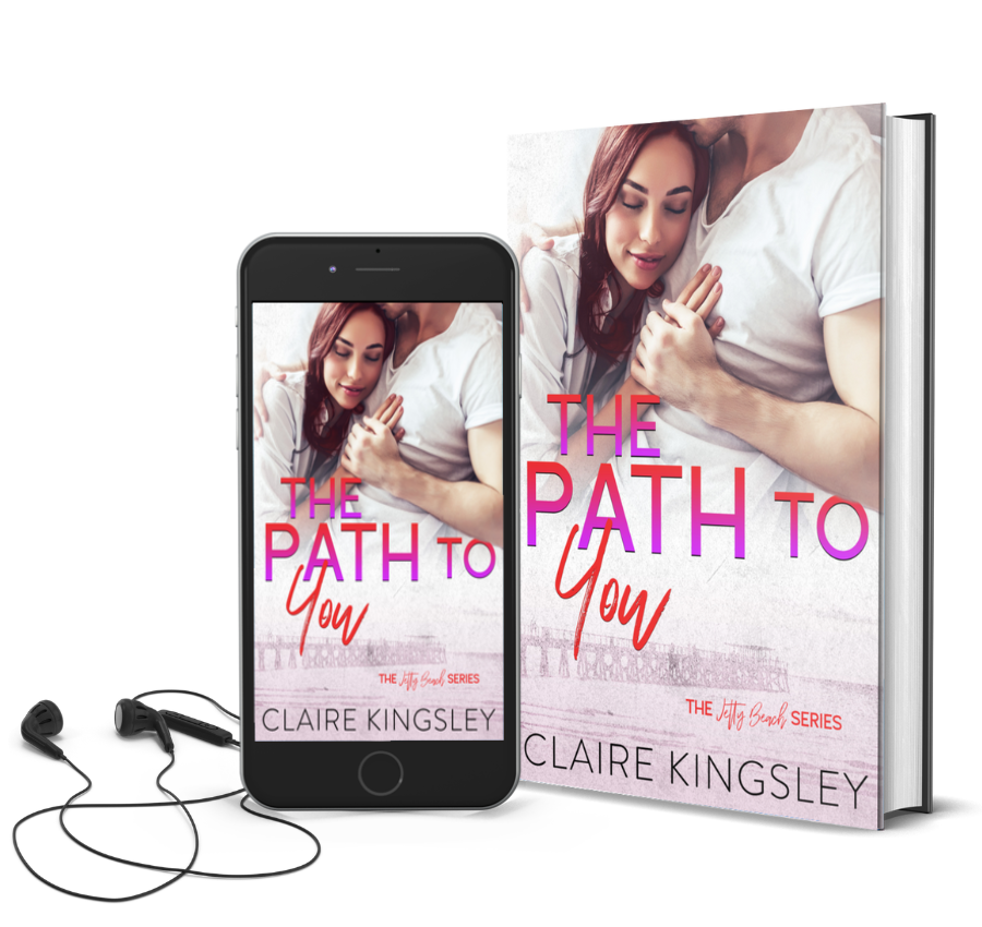 The book cover for A Path to You, a small-town romance by Claire Kingsley, is an image of a woman with red hair resting her cheek against the chest of a man while he presses his lips against the top of her head. He has one hand on top of hers as it lays gently next to her face. There are shades of red and purple. Next to the book is a phone showing the cover with a pair of wired earbuds.